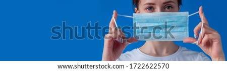 Wide banner copyspace - woman putting on medical face mask and looking at camera against blue background at home: close up front view. Self isolation, quarantine, COVID19, coronavirus, safety concept