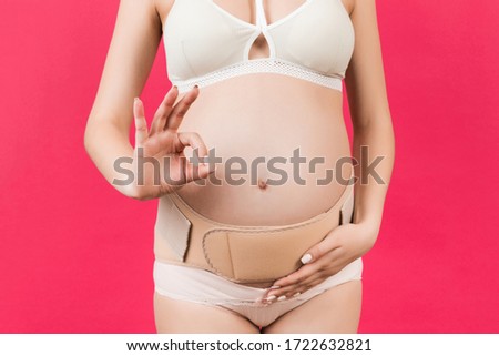 Close up of pregnant woman in underwear wearing supporting bandage and showing okay gesture at pink background with copy space. Orthopedic abdominal support belt concept.