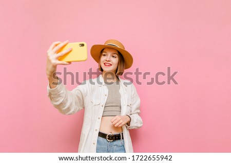 Cute girl with a smile on her face and a hat from the sun on her head, wears summer clothes, makes selfies on a pink background, looks into the camera with a happy face. Closeup lady taking selfiea