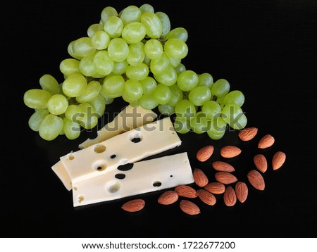 Sprigs of fresh green grapes, pieces of cheese with holes maasdam and almonds on a black background, closeup. A natural appetizer for wine. Bright picture for printing artwork