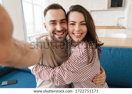 Photo closeup of young joyful couple hugging and smiling while taking selfie photo in kitchen
