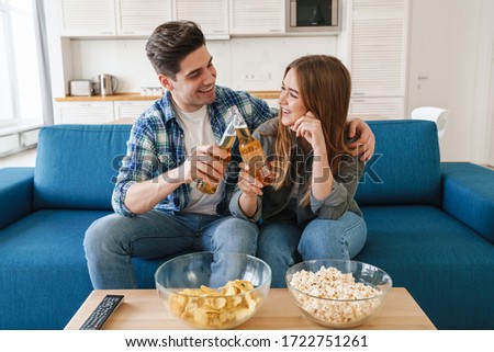 Photo of young joyful couple eating snacks and drinking bear while watching tv in modern kitchen