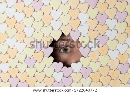 Girl looking through hole in wall of heart-shaped sweets. Dramatic shadow covers part of face. Royalty free stock photo.