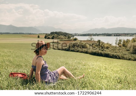 Young white woman in a blue dress and brown hat sitting on the grass from back near the lake on a background of mountains in summer.