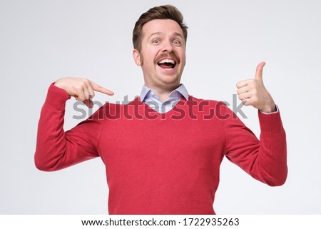 Happy bearded man in red sweater shows himself with thumbs, smiles satisfied happy, pointing oh his chest bragging. I am awesome. Studio shot Royalty-Free Stock Photo #1722935263