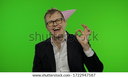 Portrait of drunk young clerk man with a hangover showing OK sign and smile. Emotions. Disheveled guy businessman in suit, festive cap, glasses. Chroma key. Office party concept