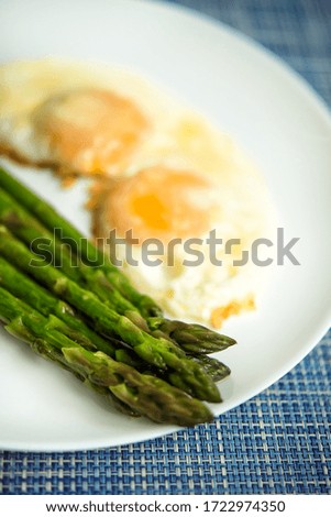 Close up picture of baked roasted fresh green asparagus with fried eggs under parmesan cheese in big white plate on blue background, top view. Seasonal spring food nutrition concept. Diet. Seasonal.