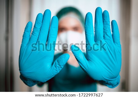 A young female doctor is blocking the entrance in her work space. Focus on hands. Prevention of Covid-19. Green uniform, stop sign with hands.