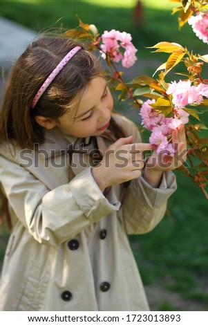 Young beautiful girl in a beige trench coat stands and enjoys the aroma of a blossoming sakura tree in the park. Photo taken in daylight.
The photo was taken with selective focus.