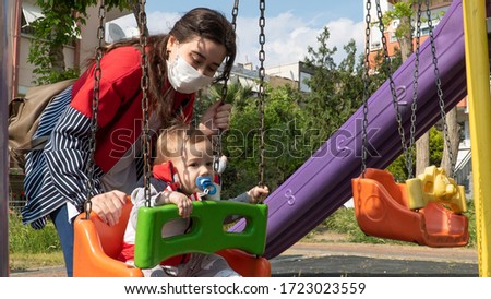 A woman in a red sweater and a white medical mask and Mom is playing with her little son swing together in the park. Family lifestyle Concept. 