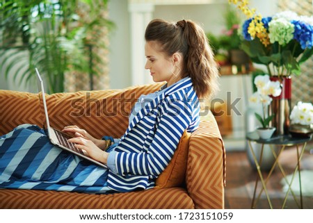 Side view of trendy housewife in blue blouse and striped jacket laying on sofa with laptop in the modern house in sunny day.