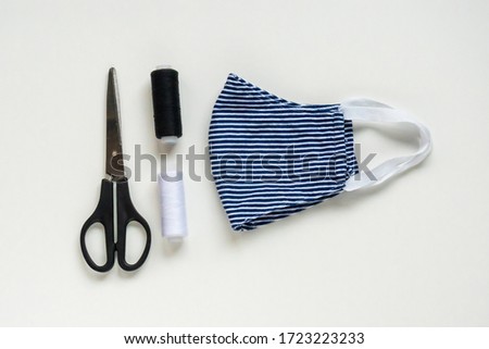 Sewing reusable and washable cotton mask during coronavirus or infection or allergy. Thread, scissors, and blue-and-white striped mask on white background. DIY, Handmade product. Royalty-Free Stock Photo #1723223233