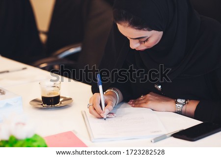 Pensive Arab woman planning working schedule writing in notebook while sitting at working place, Arabic female administrative manager making notes of information browsed on netbook.