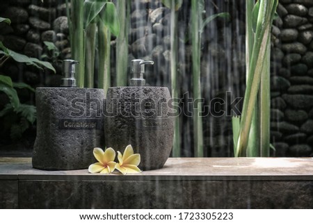 Different stone bottles with shampoo, gel and conditioner on stone shelf with frangipani  in outdoor shower. Organic cosmetics concept.