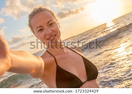 Smiling young woman taking a selfie photo at sandy beach by the sea at sunset.