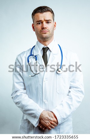 Young male doctor in a white coat over shirt with necktie, and with a stethoscope, standing and holding his hands together, looking at camera with sad face. Front portrait against white background