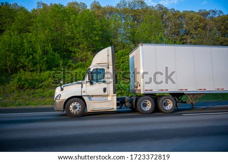 White Big rig day cab semi truck with roof spoiler for better aerodynamics and air resistance improvements transporting cargo in dry van semi trailer running on the road with green trees on the side Royalty-Free Stock Photo #1723372819