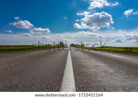 Low angle view of a tarred road receding into the distance in open countryside under a cloudy blue sky with focus to the center white line Royalty-Free Stock Photo #1723451164