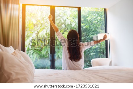 Rear view image of a woman do stretching after waking up in the morning  , looking at a beautiful nature view outside bedroom window 