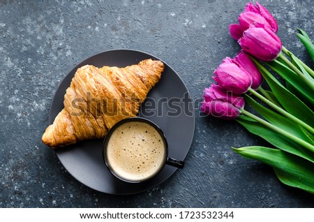 Cup of coffee with bouquet of pink tulips and croissant on black background. Valentines, Mothers, Womens Day morning concept. Valentine's day breakfast. Top view.