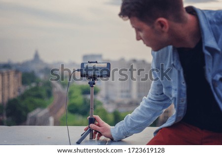 A young hipster man takes photos and videos of the cityscape at sunset from the roof on a smartphone. Smartphone on a tripod