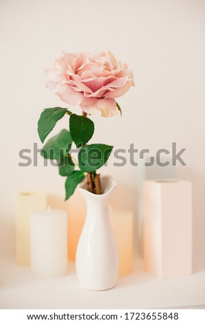 One pink rose stands in a small white ceramic vase standing next to candlesticks on a light background.