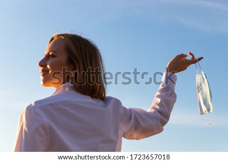 Happy woman takes off medical protective mask holds it on her finger on blue sky background, enjoys life, clean fresh air after Covid-19 pandemic, self-isolation. Quarantine is over. Soft focus Royalty-Free Stock Photo #1723657018