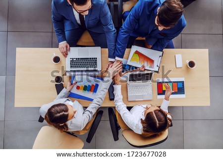 Top view of four successful business people sitting at table and stacking hands. On table are charts and statistics for stock market.