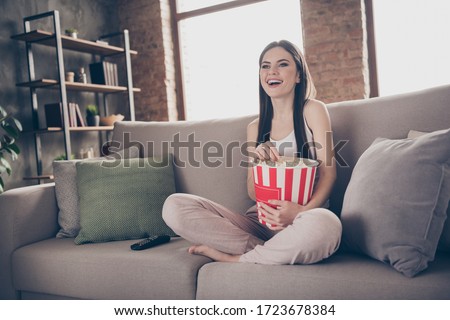 Full length photo of candid girl sit divan legs crossed watch funny movie hold box eat pop corn enjoy free time in house indoors living room