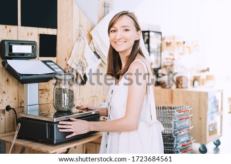 Girl weighs glass jar with hemp seeds. Woman with cotton bag chooses and buys products in zero waste shop. Weighing dry goods in plastic free grocery store. Sustainable shopping at local business Royalty-Free Stock Photo #1723684561