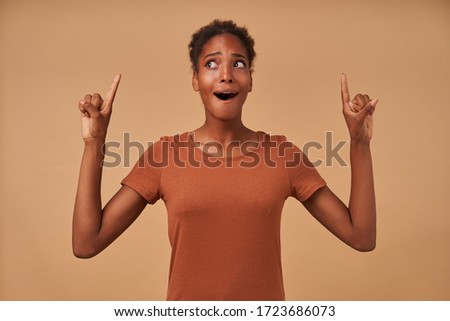 Bemused young lovely dark skinned curly woman with bun hairstyle pointing upwards with forefingers and looking excitedly aside, standing over beige background