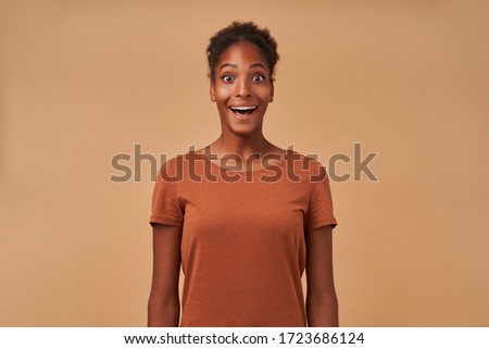 Studio shot of bemused young curly brunette female keeping her mouth wide opened while looking surprisedly at camera, isolated over beige background in casual wear