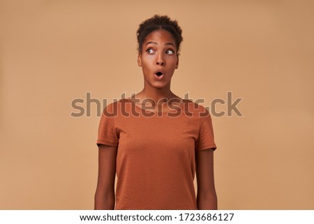 Shocked young pretty brown haired curly woman with dark skin looking amazedly aside with wide eyes and mouth opened, isolated over beige background