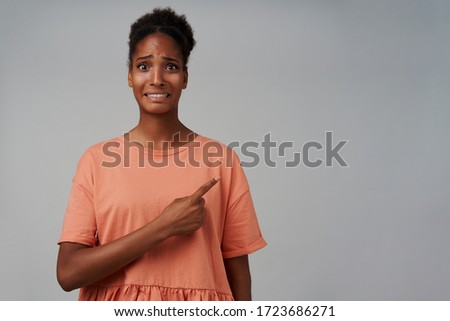 Indoor shot of confused young dark skinned curly female keeping her hand raised while showing aside with index finger and grimacing her face, standing over grey background