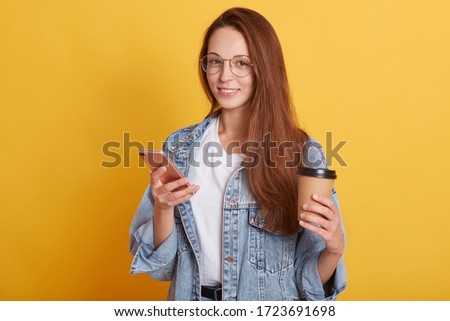 Picture of cheerful good looking young female standing isolated over yellow background in studio, holding paper cup with hot drink and smartphone, having delighted facial expression. Emotions concept.