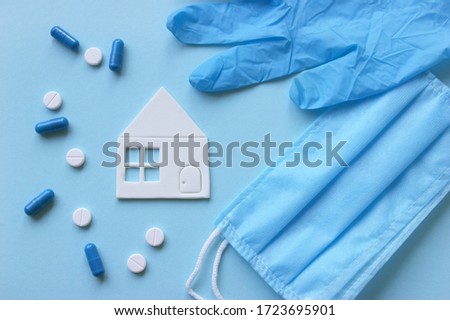 A three-layer medical mask with rubber ear straps and various medicines, tablets and in the center is a paper white house. The concept of medicine. Hope. Stay home, stay safe.