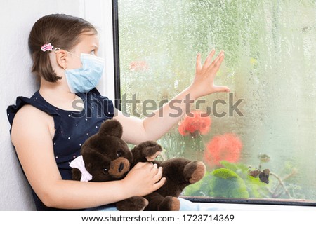 Stay at home-quarantine coronavirus pandemic prevention. Sad girl and her bunny both in protective medical masks sits on windowsill and looks out window. Prevention epidemic concept.