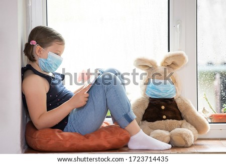 Stay at home-quarantine coronavirus pandemic prevention. Girl reading to her bunny, both in protective medical masks sits on windowsill. Prevention epidemic concept.