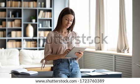Beautiful young woman using computer tablet, standing in modern living room at home, spending leisure time chatting in social network, writing message, focused blogger freelancer working online