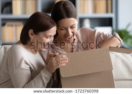 Surprised middle aged mother and adult daughter unpacking parcel together, looking in cardboard box, happy young woman with mature mum excited by delivery, received online store order Royalty-Free Stock Photo #1723760398