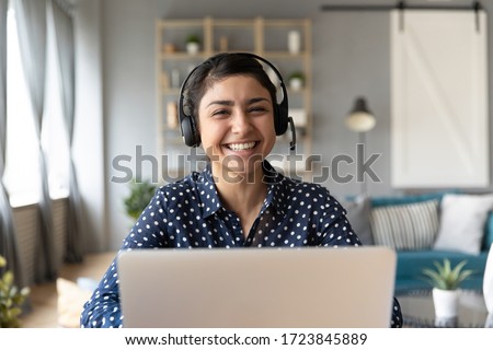Head shot cheerful smiling pretty hindu ethnic girl sitting at table with computer, wearing headphones with mic, looking at camera. Happy indian woman professional tutor educating client online. Royalty-Free Stock Photo #1723845889