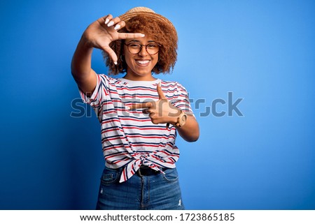African American woman with curly hair on vacation wearing summer hat and striped t-shirt smiling making frame with hands and fingers with happy face. Creativity and photography concept.