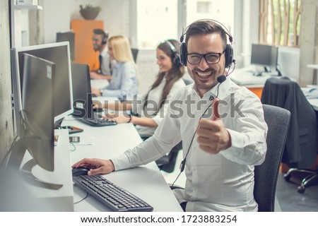 Friendly male helpline operator man with headphones showing thumb up in call center.