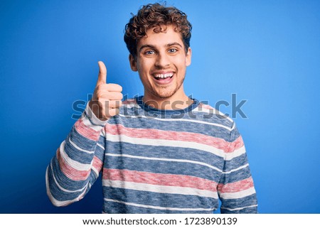 Young blond handsome man with curly hair wearing striped sweater over blue background doing happy thumbs up gesture with hand. Approving expression looking at the camera showing success.