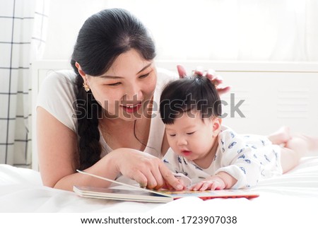 Warm moment of Asian mother playing and teaching her cute newborn baby with cartoon picture book in the bedroom at home. Storytelling, happy family, mother and baby lifestyle and pure love concept
