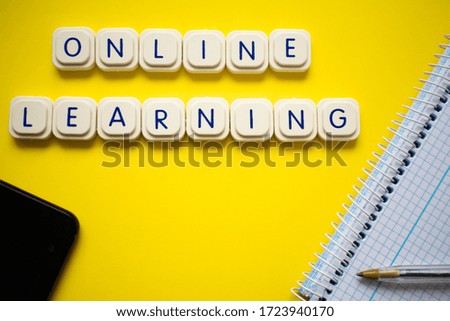 Online learning message made with game letters blocks, over a bright yellow background, with a notebook and a pen in one side and a smartphone in another. representation of learning form home.