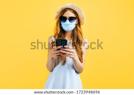 Shocked girl in a summer hat and sunglasses, wears a medical protective mask against a viral infection, uses a mobile phone on a yellow background. Quarantine, coronavirus, summer