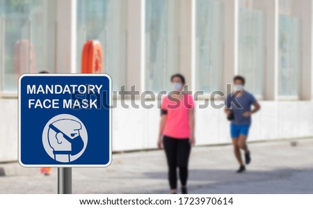 Blue sign warning of that face mask is mandatory due to Covid-19 or coronavirus on street, purposely blurred background with people training, running and wearing face mask