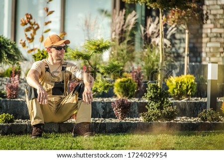Mid 30s Male Gardener Sitting Down And Relaxing On Stone Edging In Residential Backard Area.  Royalty-Free Stock Photo #1724029954