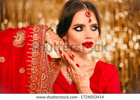 Portrait of beautiful indian girl in red bridal sari over golden bokeh. Young hindu woman model with kundan jewelry. Traditional Indian costume lehenga choli. Henna painting, mehendi on bride's hands.